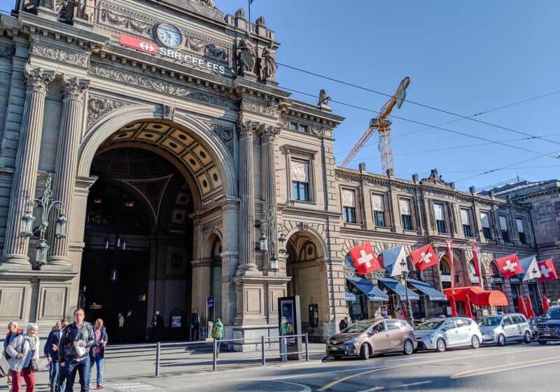 Outside of Zurich trainstation, large stone building with columns in the front, and several Swiss flags