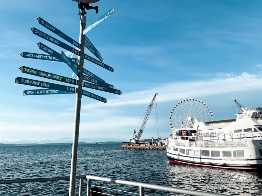 View of Seattle waterfront with a ferris wheel, ferry, and crane in the background. A directional sign pointing in a bunch of directions in the foreground