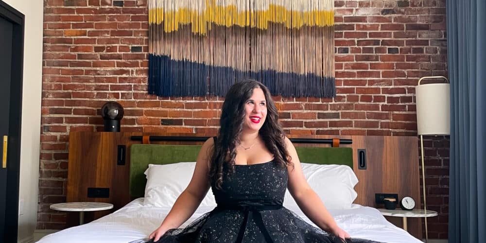 Brunette sitting on bed at Alexis Hotel in black sparkly dress