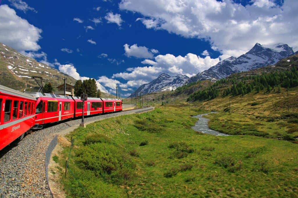 Red train with windows winding through green countryside with mountains in the background and blue sky with white clouds